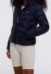 Granatowa kurtka puchowa z zamkiem błyskawicznym, kapturem i bocznymi kieszeniami. Posiada czarną metkę na piersi oraz teksturowaną tkaninę na powłoce.