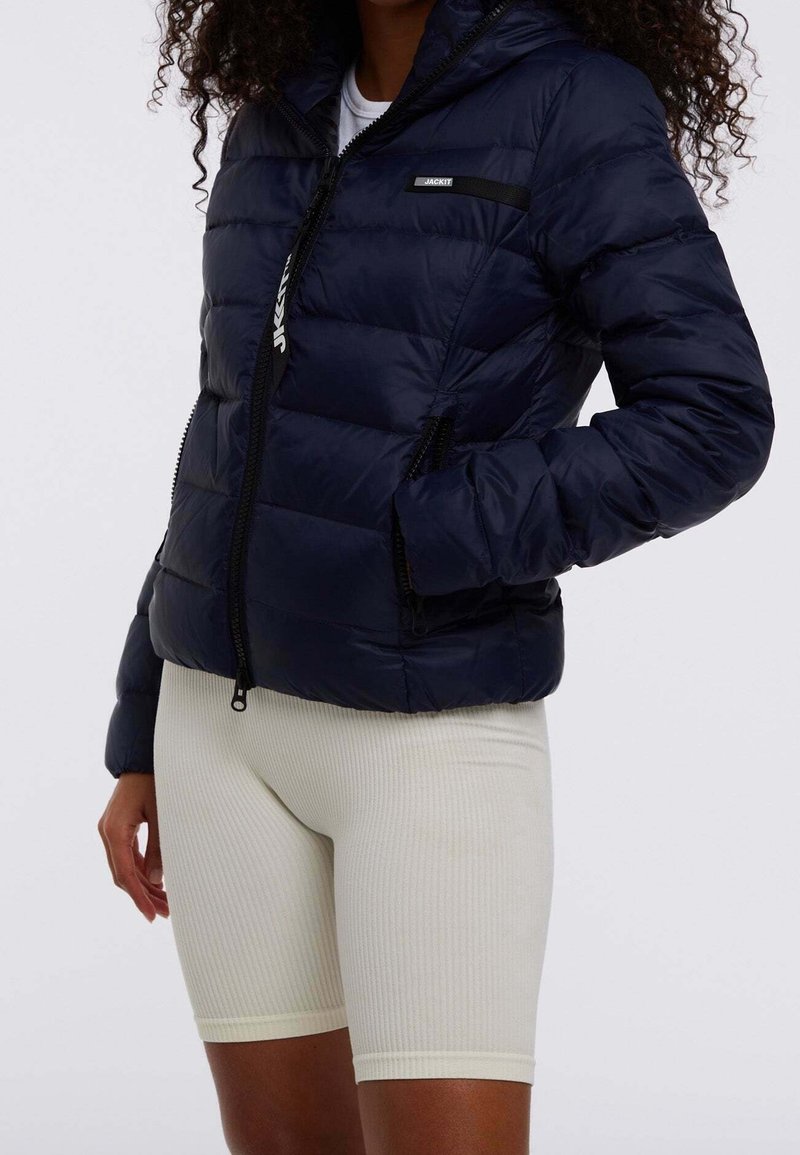Granatowa kurtka puchowa z zamkiem błyskawicznym, kapturem i bocznymi kieszeniami. Posiada czarną metkę na piersi oraz teksturowaną tkaninę na powłoce.