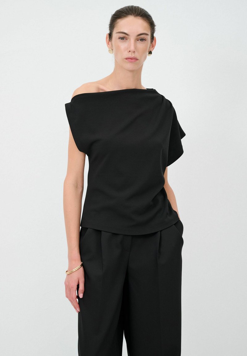 Top nero scoperto sulle spalle in tessuto liscio con design drappeggiato. Abbinato a pantaloni neri a vita alta. Stile minimalista, senza pattern né grafiche.