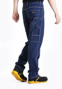 Pantalons cargo en denim bleu avec plusieurs poches, coutures contrastées et coupe décontractée. Associés à des chaussures de sport noires avec une semelle jaune.
