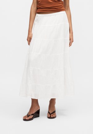 Kobieta w białej, warstwowej maxi spódnicy, brązowej bluzce bez rękawów i czarnych sandałach na koturnie, stojąca na jednolitym tle.