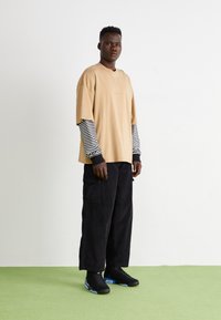Tricou oversized bej suprapus peste un maiou cu dungi negre și albe; asortat cu pantaloni cargo negri și adidași negri cu accente albastre.