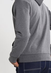 Persona che indossa un felpa grigia e pantaloni blu navy con cuciture bianche, in piedi con una mano in tasca contro uno sfondo semplice.