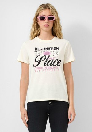 Blondynka w różowych prostokątnych okularach przeciwsłonecznych, białej koszulce z nadrukiem i czarnych dżinsach, stojąca na tle jednolitej białej ściany.