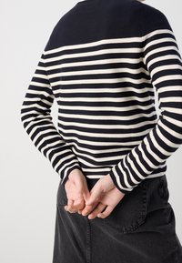 Osoba ubrana w czarno-białą w paski koszulę z długim rękawem i ciemne dżinsy, z dłońmi splecionymi za plecami, odwrócona plecami.