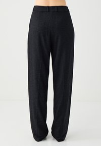 Femme portant un pantalon taille haute gris foncé ajusté, debout devant un fond blanc uni.