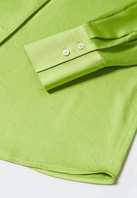 Poignet de chemise à manches longues vert clair avec deux boutons blancs, tissu lisse et coutures visibles autour de l'ourlet.