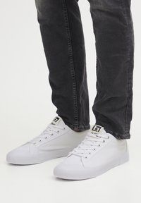 Valged kanvas tennisjalatsid madala disainiga, ovaalse varbaga, paeltega ja bränditud keelega. Kombineeritud tumesiniste slim-fit teksadega.