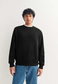 Felpa nera con scollo tondo, maniche lunghe e polsini a coste. Abbinata a jeans di denim azzurro chiaro, con vestibilità comoda.