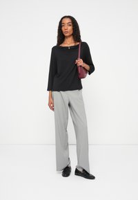 Zwarte oversized top met een bootnek, grijs gestreepte broek met wijde pijpen en zwarte schoenen. Model houdt een bordeauxrode schoudertas vast.