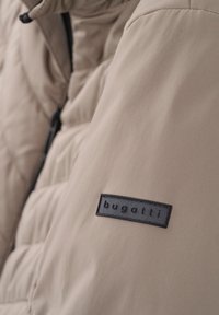 Beige gepolsterte Jacke mit gestepptem Muster, ausgestattet mit einem schwarzen rechteckigen Etikett mit dem eingeprägten Schriftzug "bugatti" am Arm.