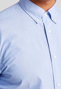 Himmelblaues Hemd mit Knöpfen aus strukturiertem Stoff, mit einem klassischen Kragen und weißen Knöpfen, das eine glatte Oberfläche betont.