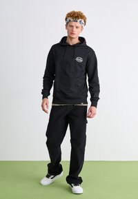 Sudadera negra con capucha y bolsillo frontal, combinada con pantalones negros de corte holgado. El modelo lleva zapatillas blancas con detalles en negro.
