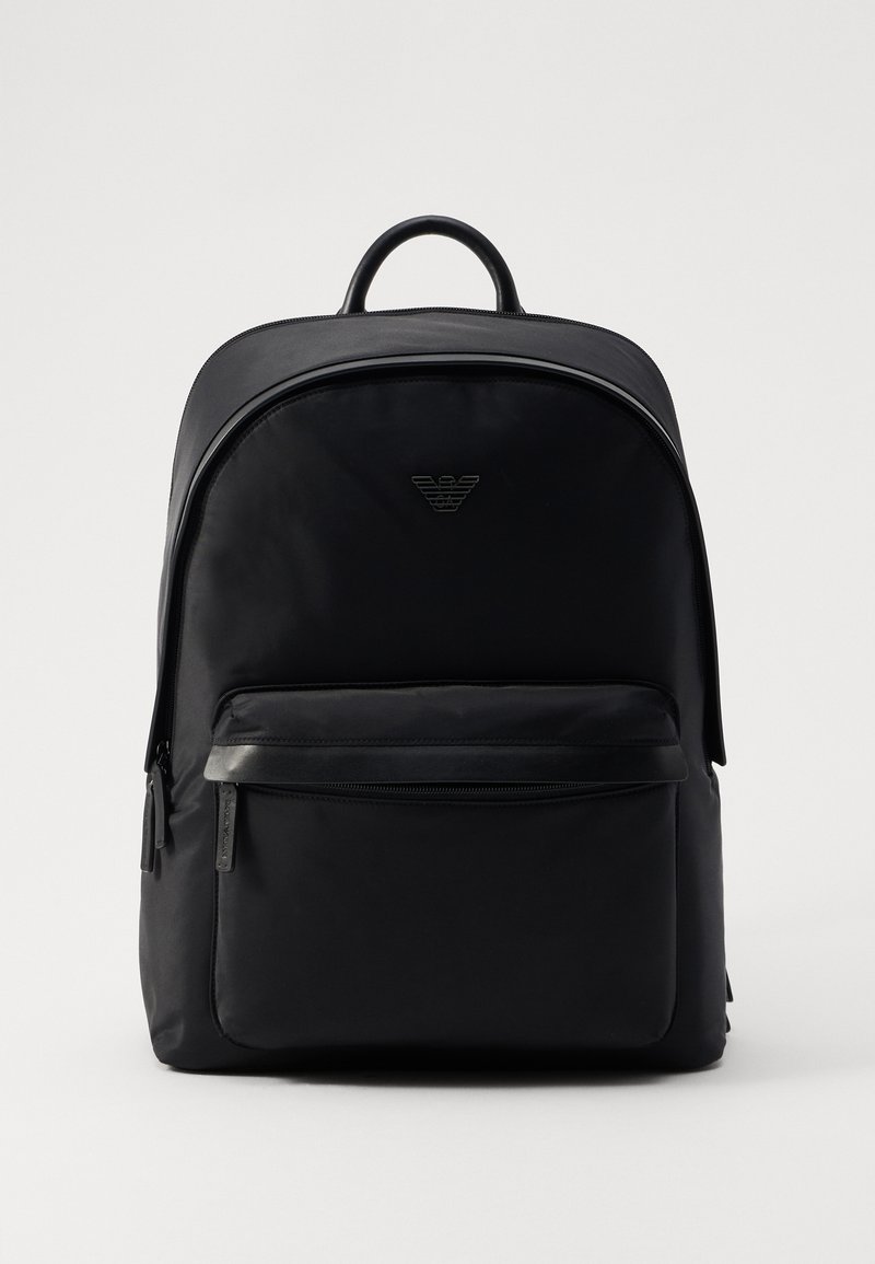 Mochila de nylon negra con forma redondeada, que cuenta con un bolsillo frontal con cremallera, correas ajustables y un logo sutil embosado en la parte frontal.