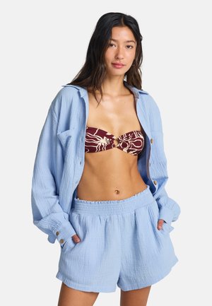 Lichtblauw, overhemd met gerimpelde textuur en oversized pasvorm met opgerolde mouwen, gecombineerd met bijpassende shorts. Een bordeauxrood- en crèmekleurige bandeau met bloemmotief eronder.