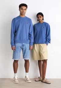 Sudaderas de algodón azul, pantalones cortos de mezclilla claros, pantalones cortos color crema y zapatos beige. Las sudaderas presentan un pequeño logo; ajuste relajado, puños acanalados.