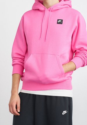 Persoon draagt een felroze Nike Air hoodie met voorzak en zwarte Nike trainingsbroek, staat met één hand in de zak.