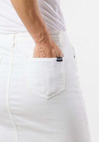 Jupe en denim blanc au design ajusté, avec une poche arrière arborant une étiquette noire et une texture subtile. Main reposant dans la poche.