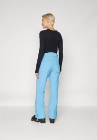 Pantaloni de schi blue, cu talie înaltă și fermoare laterale, asortați cu un top negru cu mâneci lungi. Pantalonii sunt fabricați dintr-un material neted și flexibil.