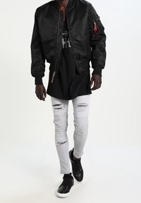 Veste bomber noire avec poignets côtelés, long t-shirt noir en dessous, jean skinny blanc déchiré, et baskets noires à enfiler.