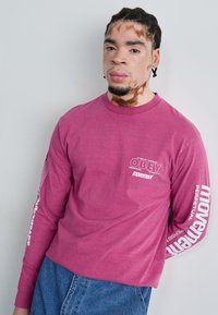 Camisa de algodón rosa de manga larga y cuello redondo, con estampados gráficos blancos en las mangas y el pecho, de corte relajado, combinada con vaqueros de denim azul.