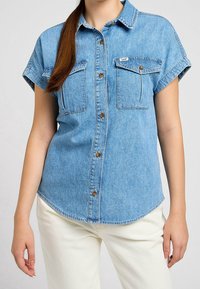 Light blue denim short-sleeve shirt with button closure, two front pockets, and a rounded hem. Features a classic collar and subtle stitching details.