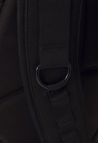 Black fabric bag detail showing webbing strap, metal D-ring, and a textured pattern on the surface. The material appears durable and rugged.