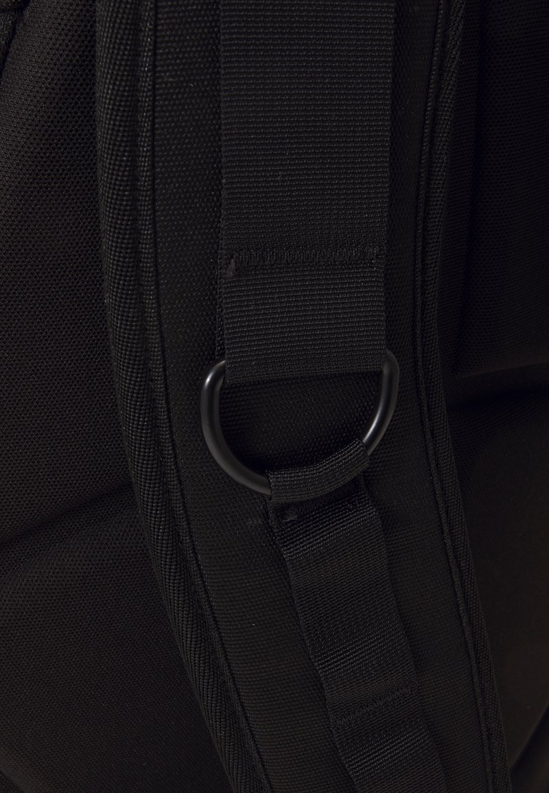 Black fabric bag detail showing webbing strap, metal D-ring, and a textured pattern on the surface. The material appears durable and rugged.