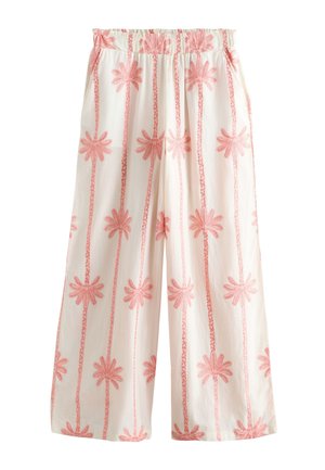 Kvinde iført hvide bukser med vide ben og matchende ærmeløs crop top med pink palmeprint, står på sandet jord nær et træhegn.