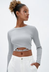 Top corto de manga larga gris con canalé y un corte circular en la cintura, adornado con herrajes de cadena plateada. Combinado con pantalones blancos de talle alto.