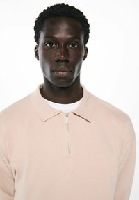 Hellrosa Sweatshirt-Polo mit Reißverschluss, standardmäßigem Kragen, strukturiertem Stoff und einer weißen Unterlage, die am Ausschnitt sichtbar ist.