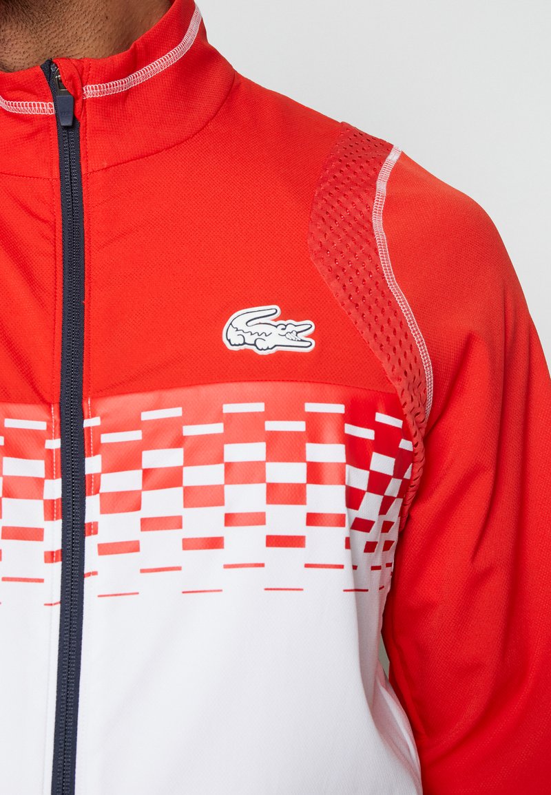 Giacca sportiva rossa con colletto alto, caratterizzata da un motivo a scacchi bianchi sul petto e un piccolo logo bianco sul lato sinistro.