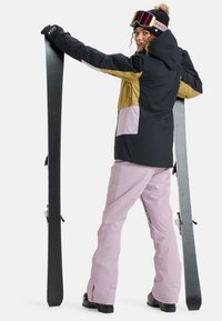 Skiausrüstung bestehend aus einer schwarz-goldenen Jacke mit pinken Akzenten, kombiniert mit hellpinken Hosen. Der Skifahrer hält zwei schwarze Skier und trägt eine Mütze sowie eine Skibrille.