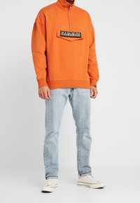 Orange zip-up sweatshirt, featuring a front pocket with a logo patch, paired with light blue jeans and beige sneakers. Smooth texture, relaxed fit.