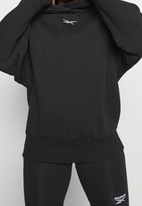 Sweatshirt à capuche noir avec une coupe décontractée et un ourlet côtelé. Présente un petit logo sur la poitrine et des manches longues. La texture semble douce.