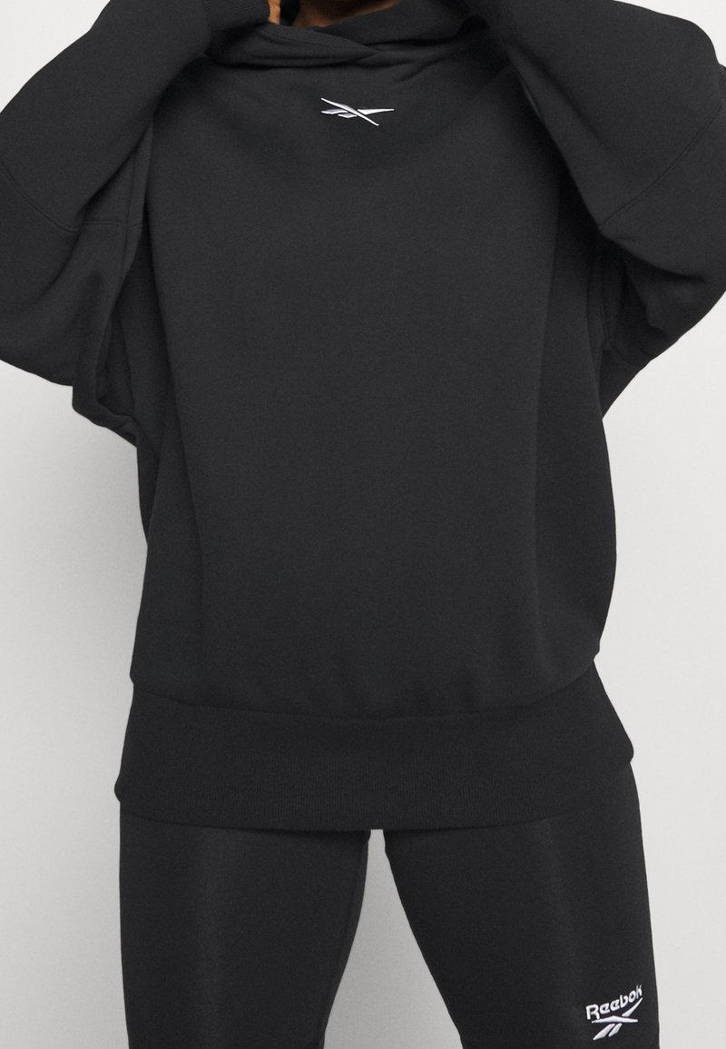 Sweatshirt à capuche noir avec une coupe décontractée et un ourlet côtelé. Présente un petit logo sur la poitrine et des manches longues. La texture semble douce.