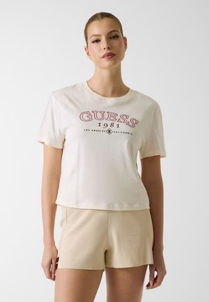 Giovane donna con indosso una maglietta bianca Guess 1981 Los Angeles California e pantaloncini beige, in piedi contro uno sfondo chiaro uniforme.