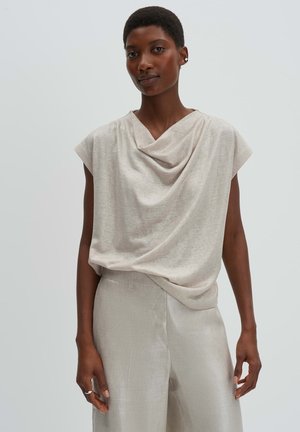 Donna con capelli corti che indossa una canottiera drappeggiata beige e pantaloni a vita alta abbinati, su uno sfondo grigio chiaro uniforme.