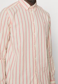 Camisa de botones clara con rayas rojas verticales, cuello estándar, mangas largas y pequeño logo bordado en el pecho.