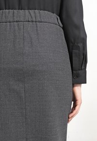 Jupe crayon en tissu gris avec une taille élastique, surface texturée et subtile brillance, associée à un blouse noire à manches longues.