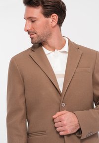 Bruine wollen blazer met een gladde textuur, revers, sluiting met één knop en een borstzak. Gecombineerd met een witte overhemd met kraag.