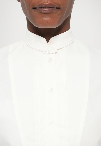 Biely košielový dres s výrazným golierom, s jemnými plisovanými záhybmi a predným zapínaním na gombíky. Hladká textúra a ľahký materiál.