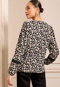 Blusa nera floreale con margherite bianche e rosa, maniche lunghe a palloncino, dettagli in pizzo ai polsini e tessuto morbido e strutturato.