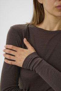 Woman with light skin wearing a brown long-sleeve top, crossing her left arm and touching her right shoulder against a plain background.