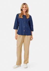 Camicia in denim blu scuro, con maniche a tre quarti, abbinata a pantaloni beige larghi e sneakers bianche.