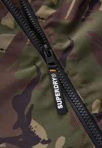 Kamuflāžas jaka, fabricēta zaļās un brūnās krāsās, ar melnu rāvējslēdzēju, redzamu logotipa etiketi un kontrastējošu šuvju apdari.