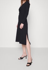 Robe noire à manches longues avec un décolleté en V, corsage ajusté et jupe avec fentes latérales. Portée avec des mules à talons ouverts noires.