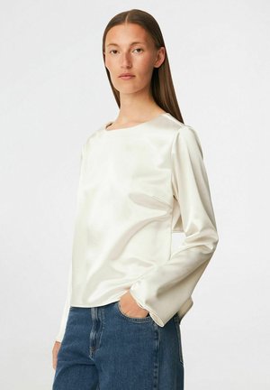 Kvinde med langt brunt hår iført en hvid satin langærmet top og blå jeans med den ene hånd i lommen mod en ensfarvet baggrund.