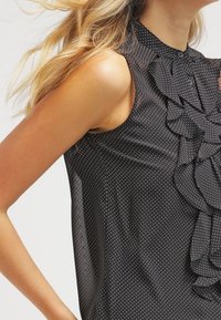 Blusa negra sin mangas con lunares blancos, que presenta un frente con volantes y un collar abotonado, llevada por una persona con cabello claro.