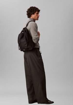 Uomo che indossa una camicia grigia a maniche lunghe e pantaloni larghi scuri, con uno zaino nero, in piedi su uno sfondo uniforme grigio chiaro.